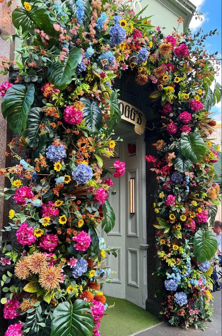 Porte d'entrée vert amande encadrée d'une arche foisonnante d'hortensias fuchsia et bleus, de tournesols et de feuilles de monstera