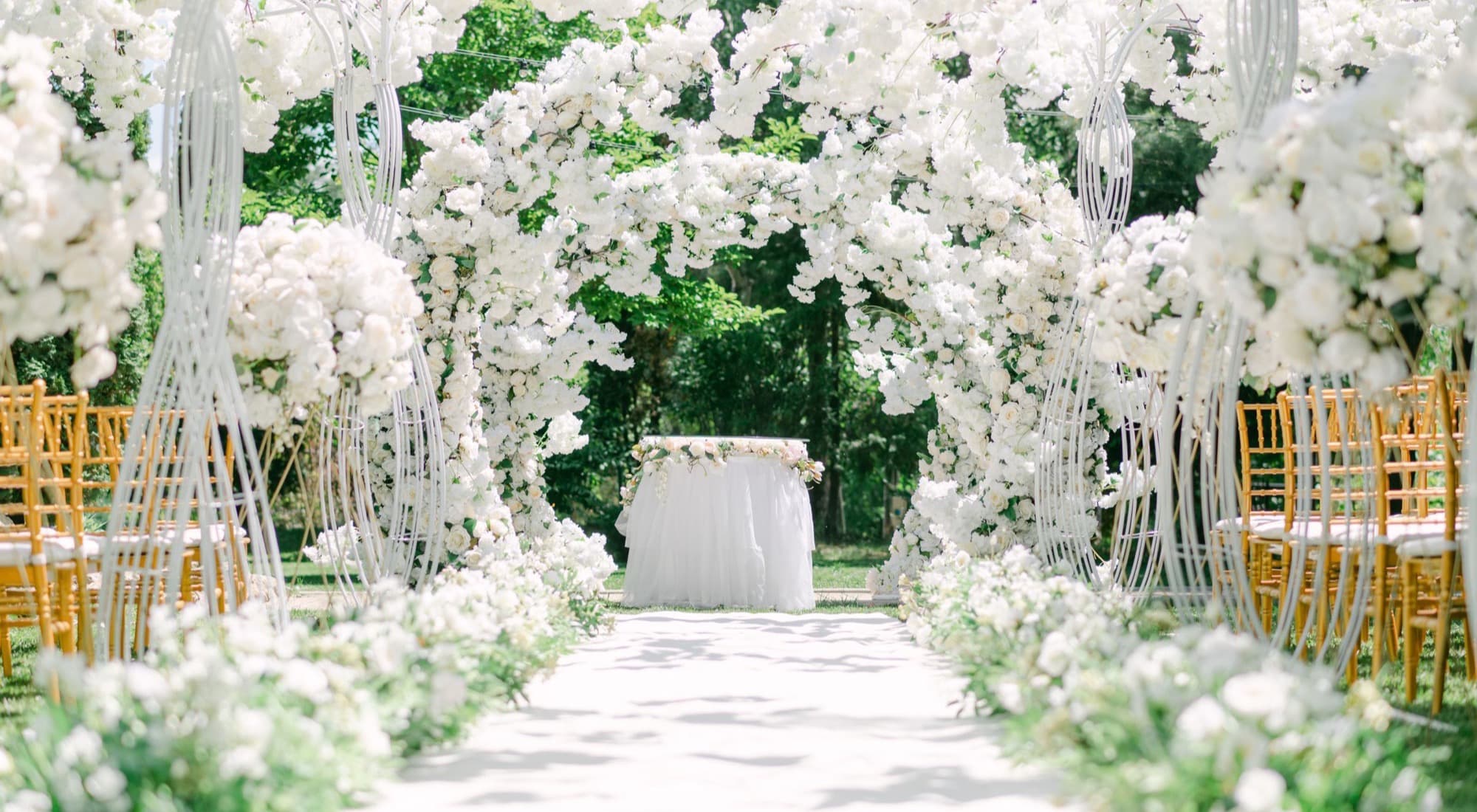Allée de cérémonie bordée d'arches de roses blanches, entre deux rangées de chaises dorées